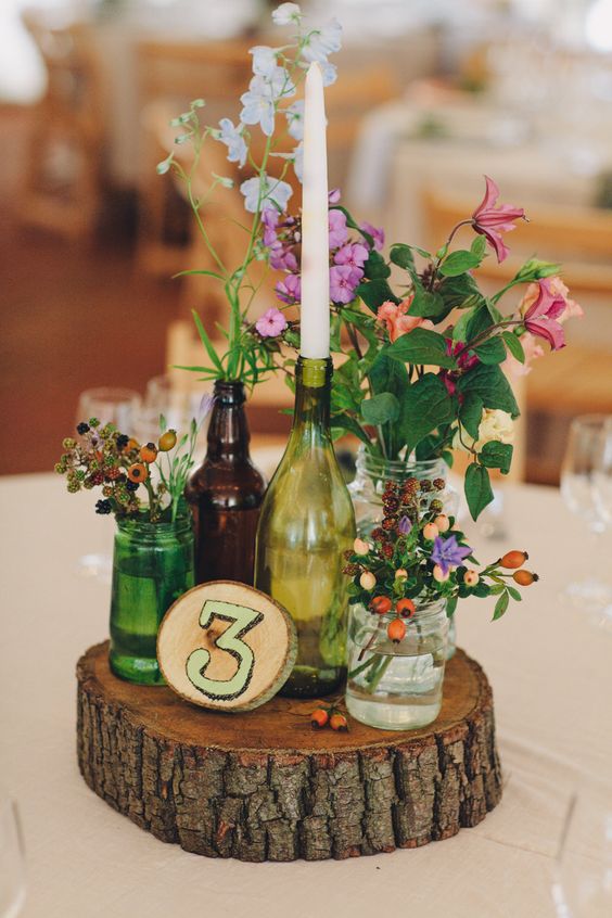 Brown and green bottles on slabs of wooden tree stumps as table centre pieces with glass jars filled with wild flowers – Image by LM Weddings Photography